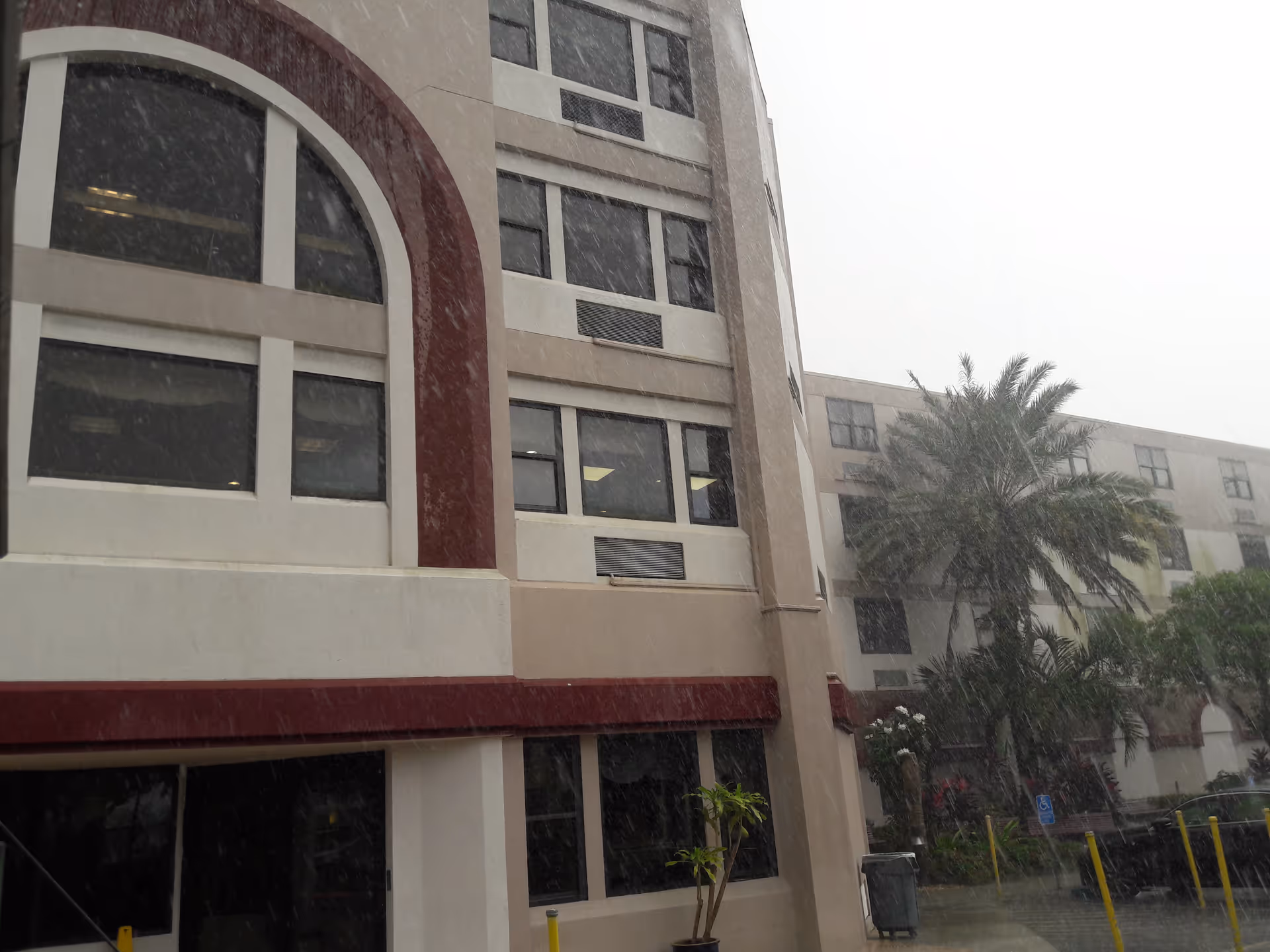 Front exterior of a multi-story nursing facility with arches, windows, palm tree in rain