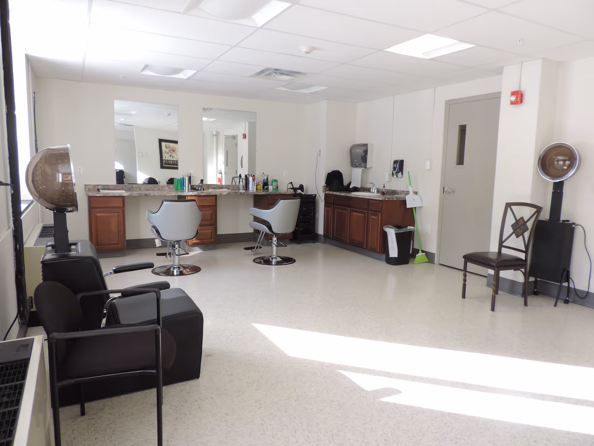 A bright, clean salon room with two styling chairs in front of a long counter with mirrors. There are hair care products on the counter, two hair dryers on stands, a black chair, a brown chair, a sink with cabinets, a trash bin, and cleaning supplies in the corner.