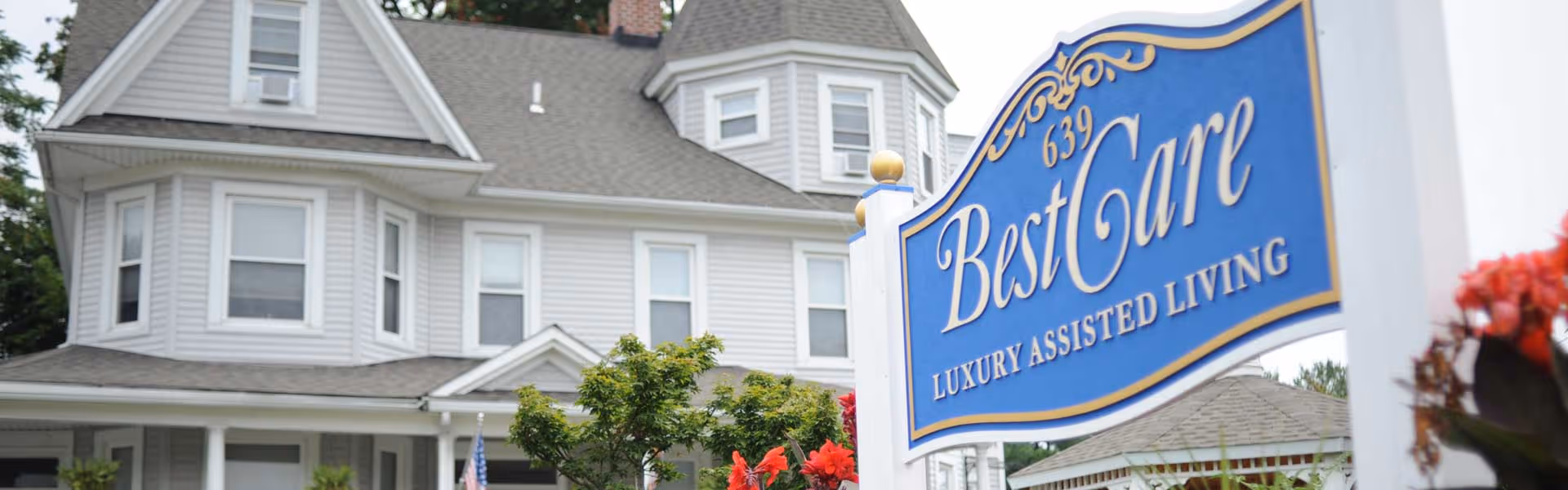 Exterior view of a large, multi-story white house with multiple windows and a porch, featuring a blue and gold sign in the foreground that reads '639 BestCare Luxury Assisted Living'.