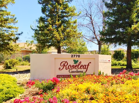 Front exterior of Roseleaf Gardens sign, surrounded by colorful flower beds and trees.