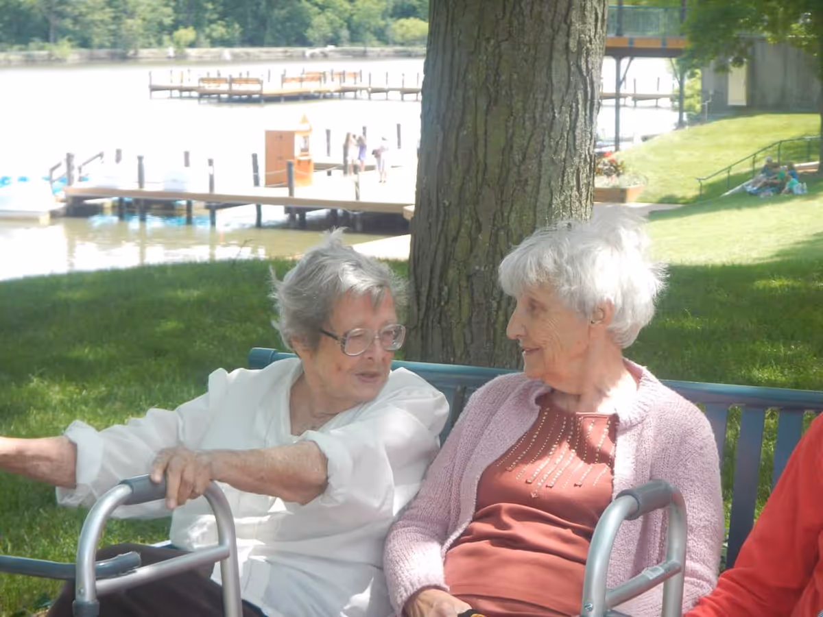 Two elderly women sitting on a bench outdoors near a tree, engaged in conversation. One woman is wearing glasses and a white shirt, while the other is wearing a pink cardigan and a red top. In the background, there is a dock extending into a body of water with a few people walking on it, and a grassy area with other people sitting on the lawn.