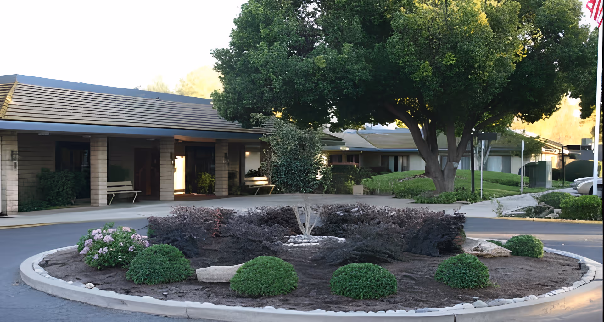 Driveway with a landscaped circular island and a large tree in front of a single-story assisted living building entrance with benches.