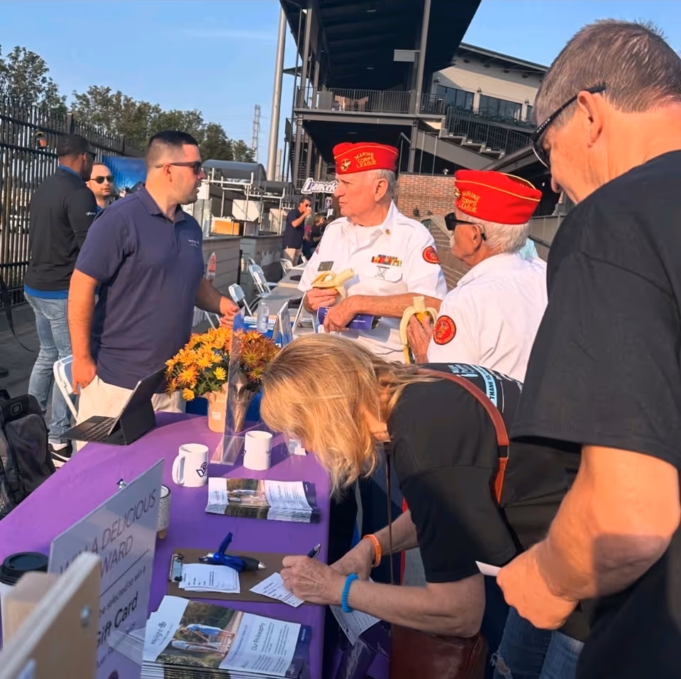 Outdoor event with veterans in red caps and volunteers at a purple table distributing brochures.