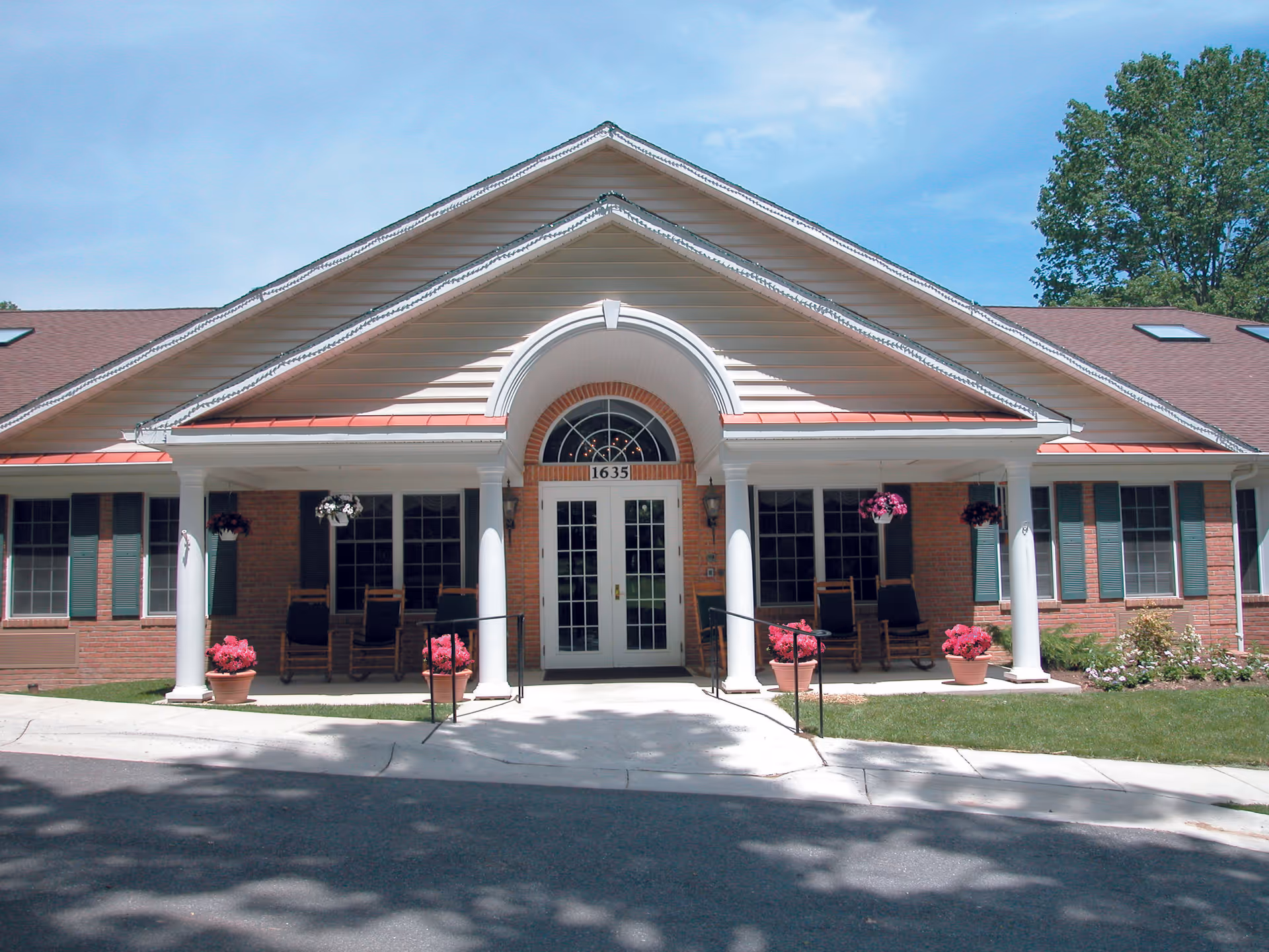 A brick senior living facility front entrance with white columns, hanging baskets, and potted flowers.