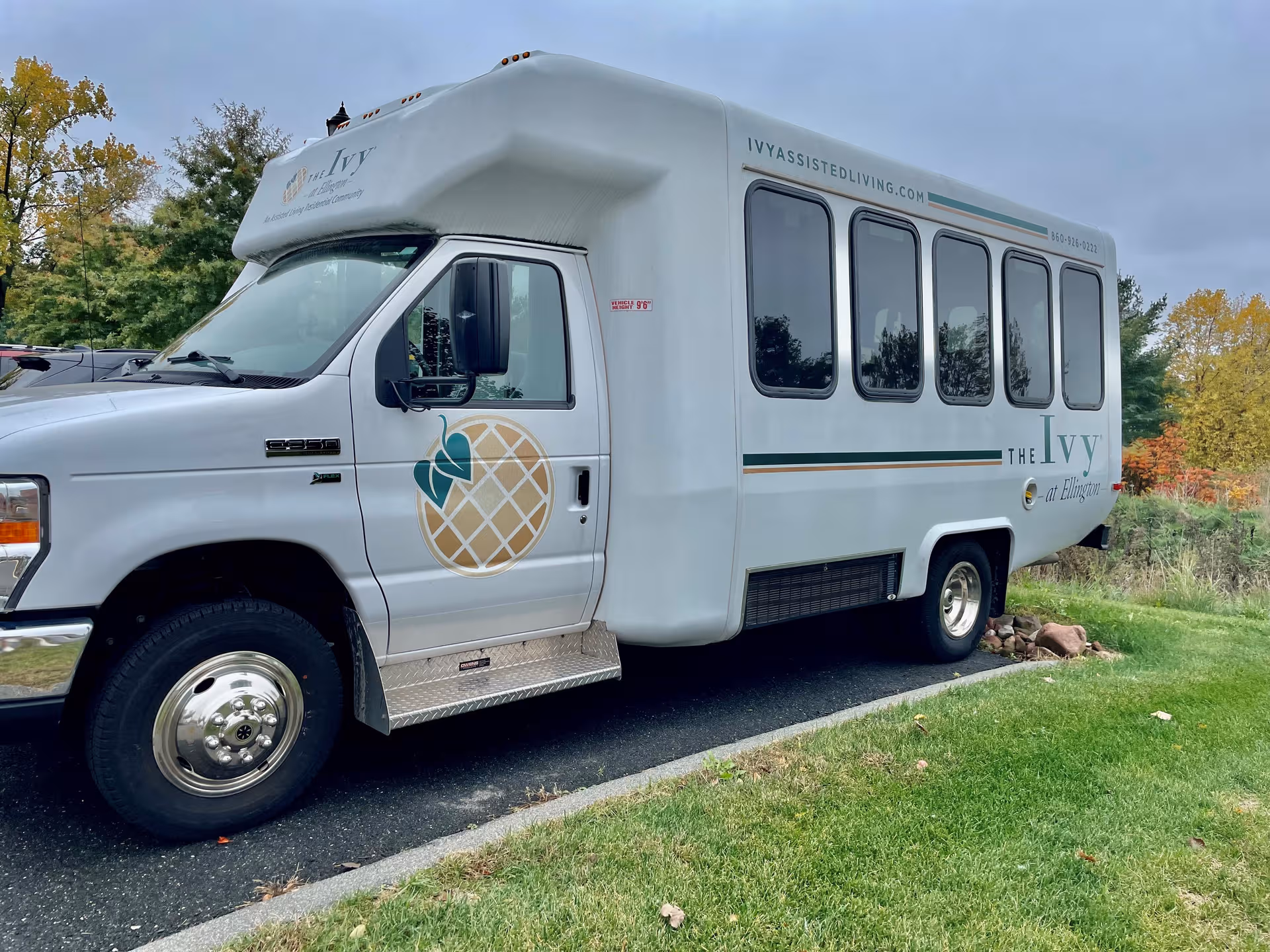 White shuttle bus branded The Ivy at Ellington parked on a paved driveway beside trees.