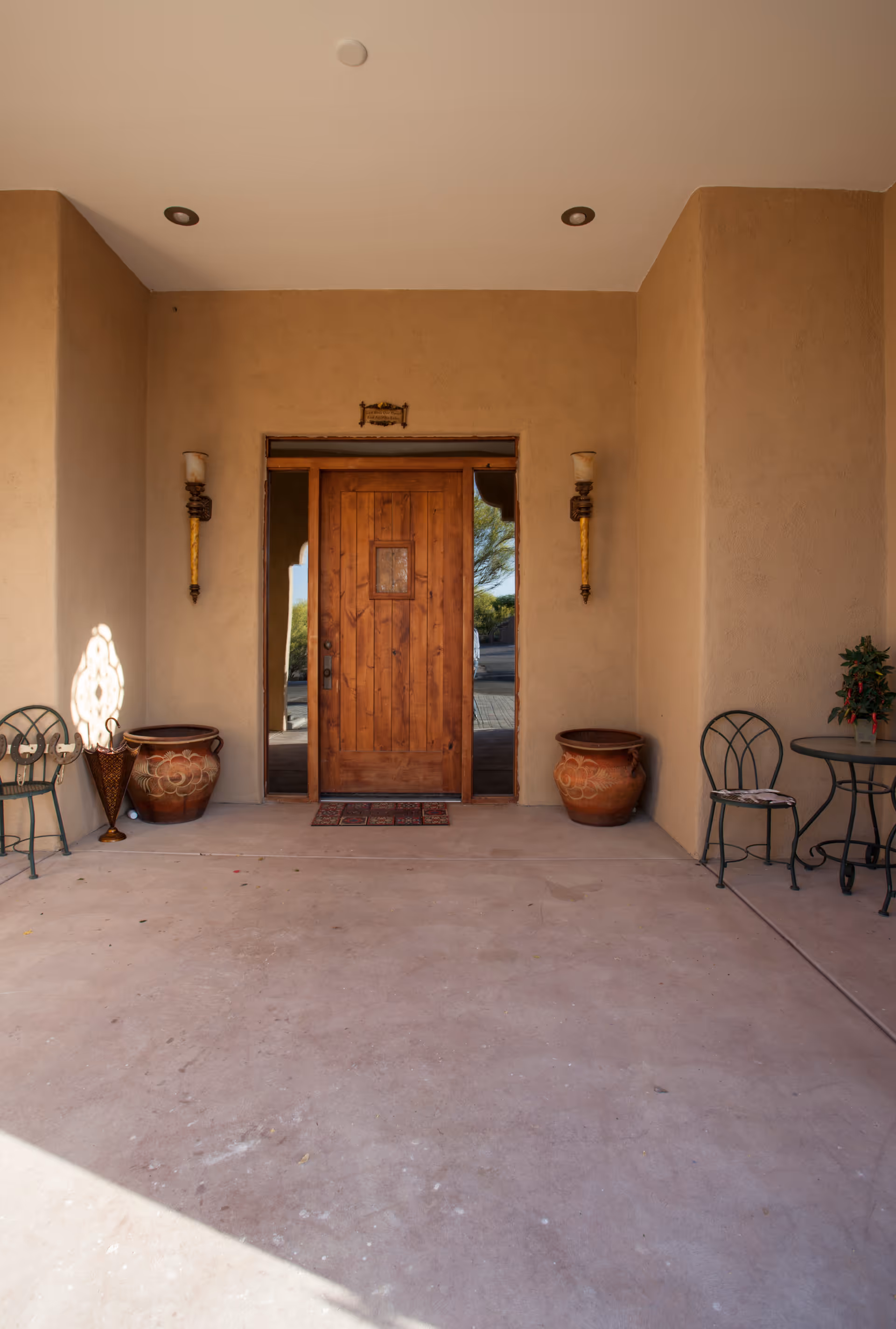 A covered entrance area with a wooden front door centered on a beige stucco wall. On either side of the door are two large decorative clay pots. Two wall-mounted sconces with candles flank the door. To the left, there is a metal chair and a decorative umbrella stand. To the right, there is a small round metal table with a chair and a small potted plant on the table. The floor is a smooth concrete surface.