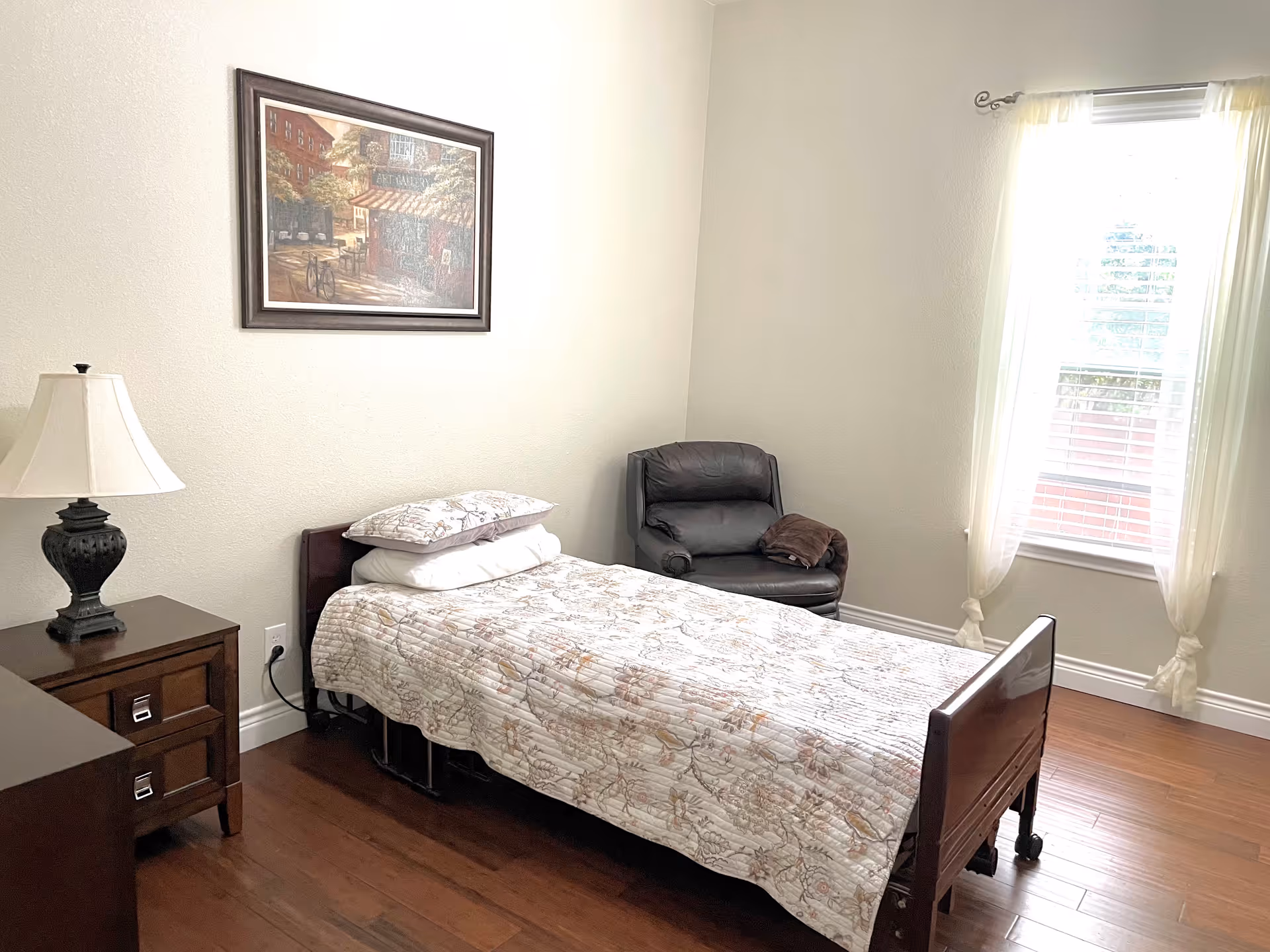 A small bedroom with a single bed covered with a floral quilt and two pillows. Next to the bed is a wooden nightstand with a decorative lamp. A framed painting hangs on the wall above the bed. There is a black armchair with a brown blanket draped over one arm in the corner near a window with sheer white curtains. The room has wooden flooring and light-colored walls.