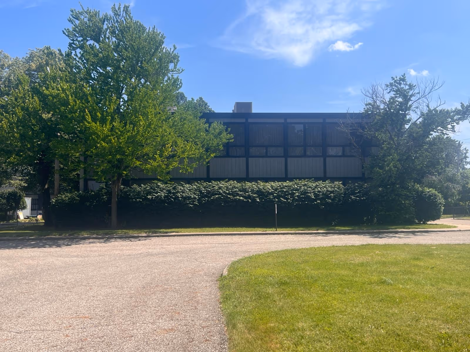 Two-story institutional building façade partly obscured by trees and shrubs under a blue sky.