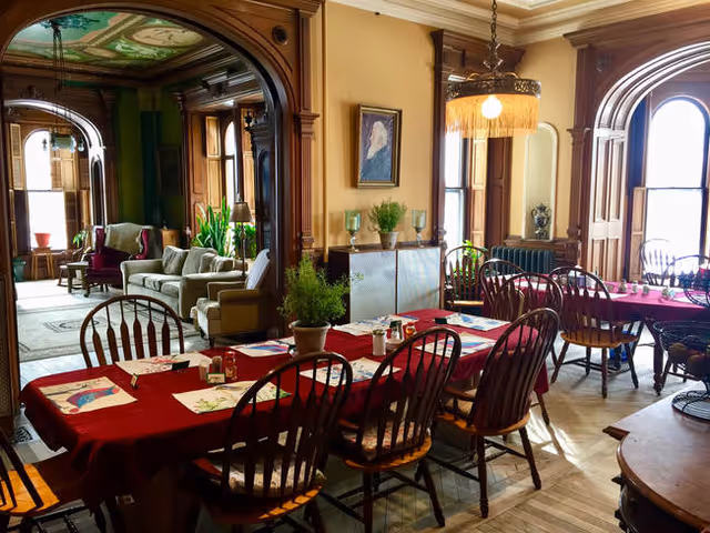 Sunlit dining room with long tables covered in red tablecloths, wooden chairs, potted plants, and an adjacent wood-paneled sitting area.