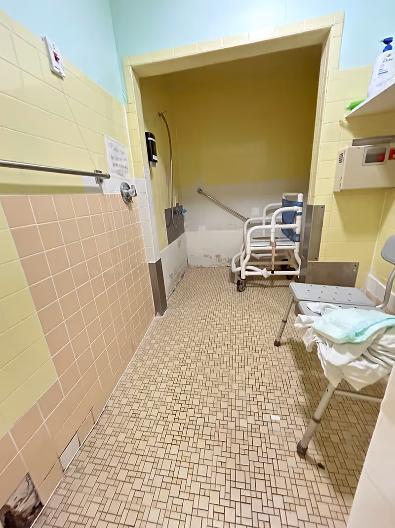 Narrow tiled bathroom with walk-in shower, wheeled chair, and medical supplies.