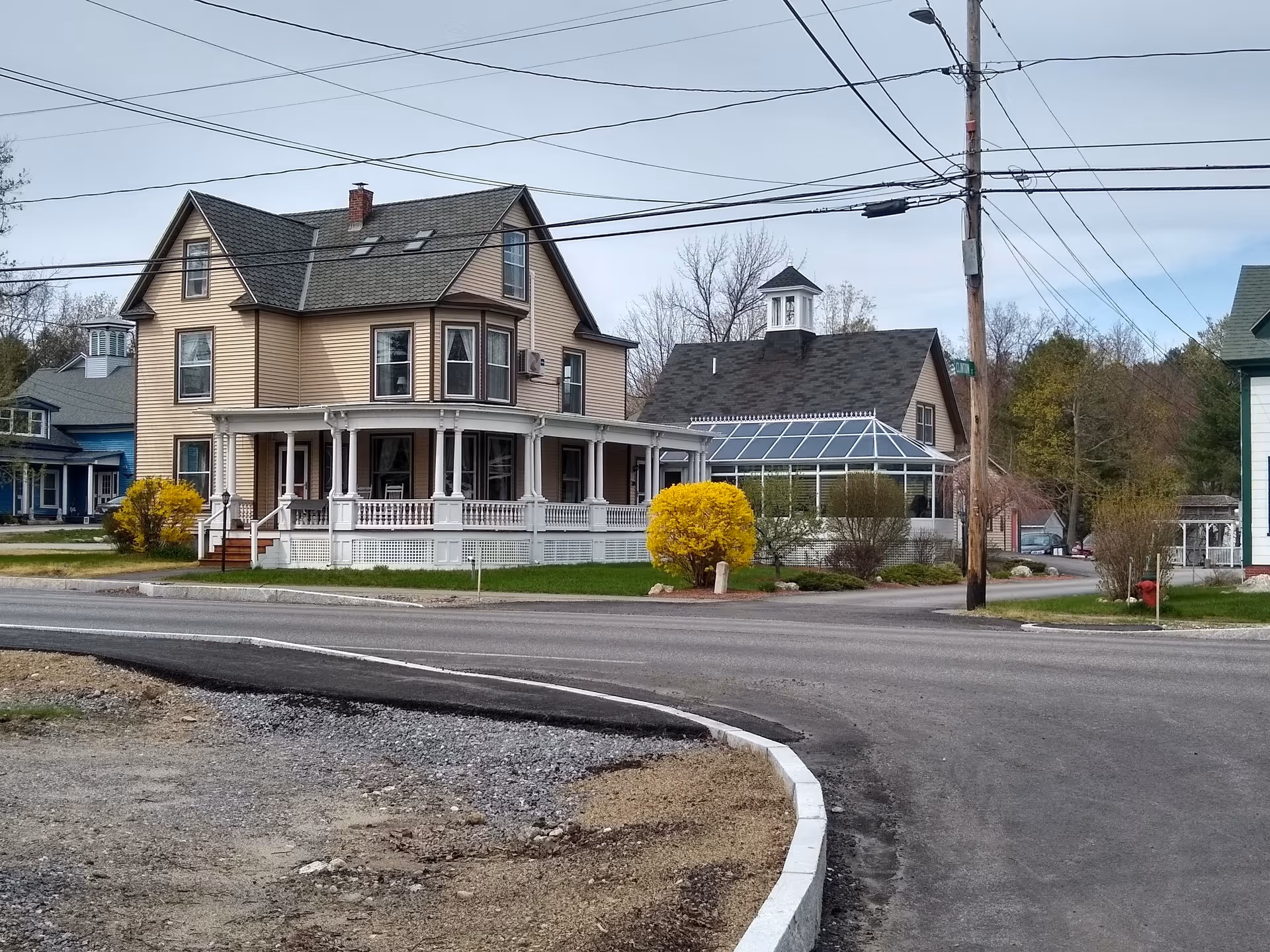 Beige Victorian-style house with a wraparound porch and adjacent glass sunroom beside a street and utility poles.