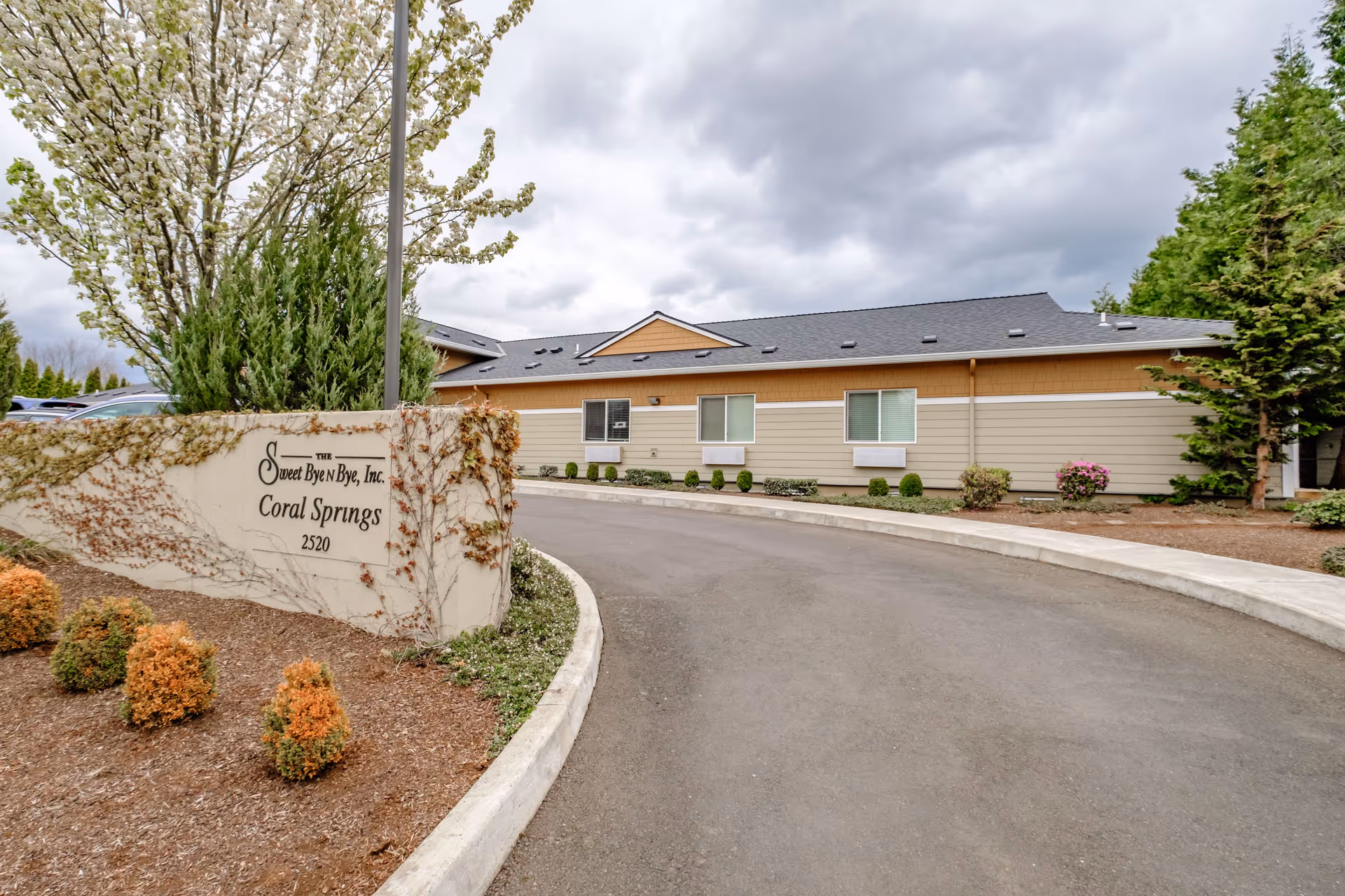 Exterior view of a single-story building with beige and light brown siding, surrounded by landscaped bushes and trees under a cloudy sky. A curved driveway leads to the building. A sign in the foreground reads 'The Sweet Byen Bye, Inc. Coral Springs 2520'.