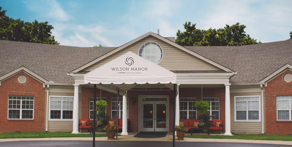 Front entrance of Wilson Manor senior living facility with a white canopy, columns, and brick facade.