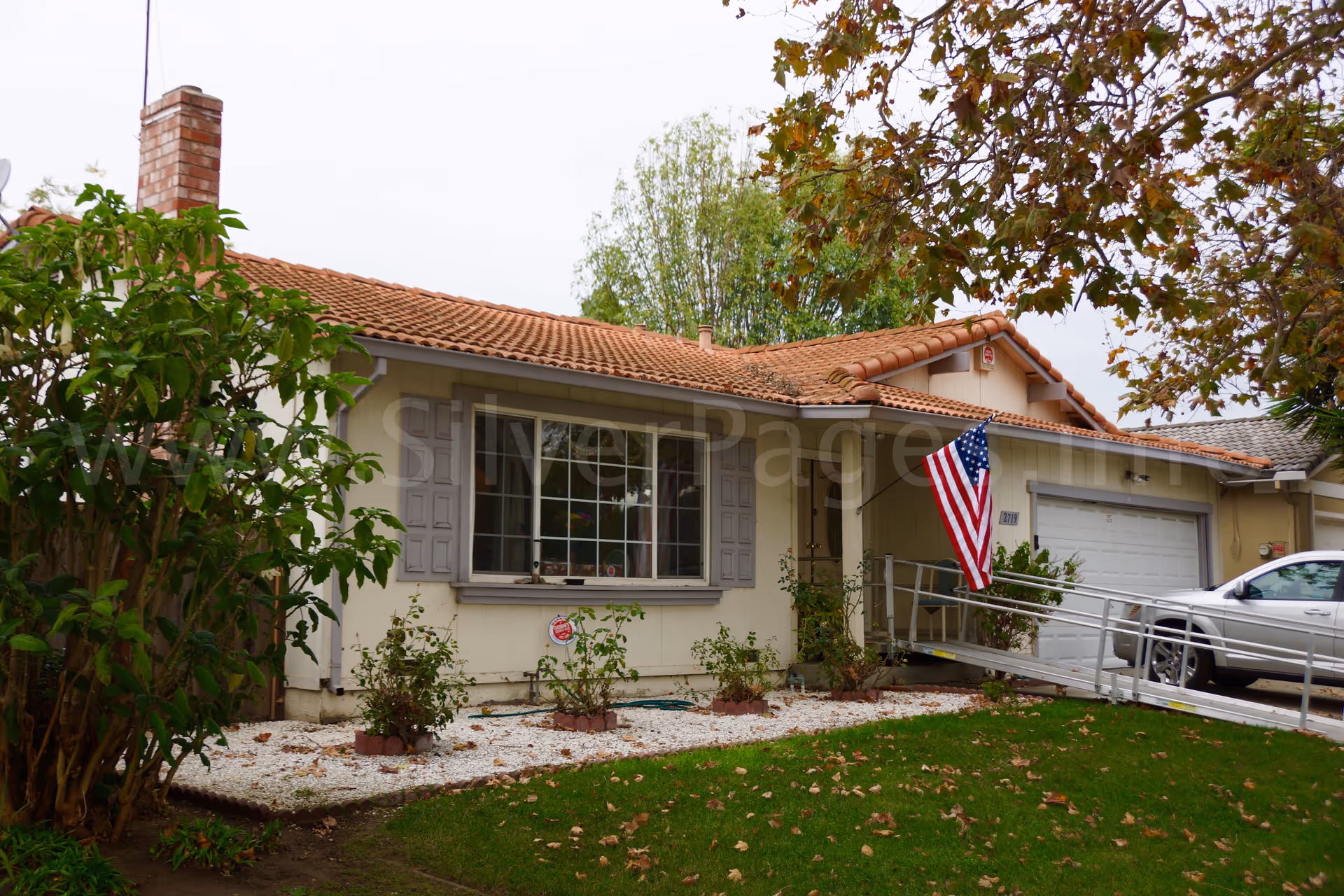 Single-story beige care facility with a ramp, orange-tiled roof, and American flag at the front