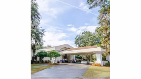 Exterior view of Surrey Place Nursing Center showing the entrance with a covered driveway, surrounded by trees and greenery under a partly cloudy sky.