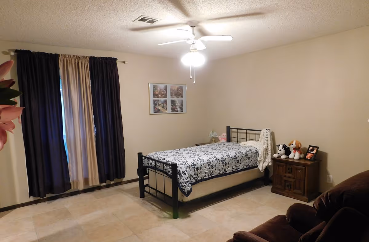 A simple bedroom with a single bed covered with a floral patterned blanket, a wooden nightstand with stuffed animals and a framed photo, a ceiling fan with lights, a window with dark and light curtains, and a brown recliner chair.