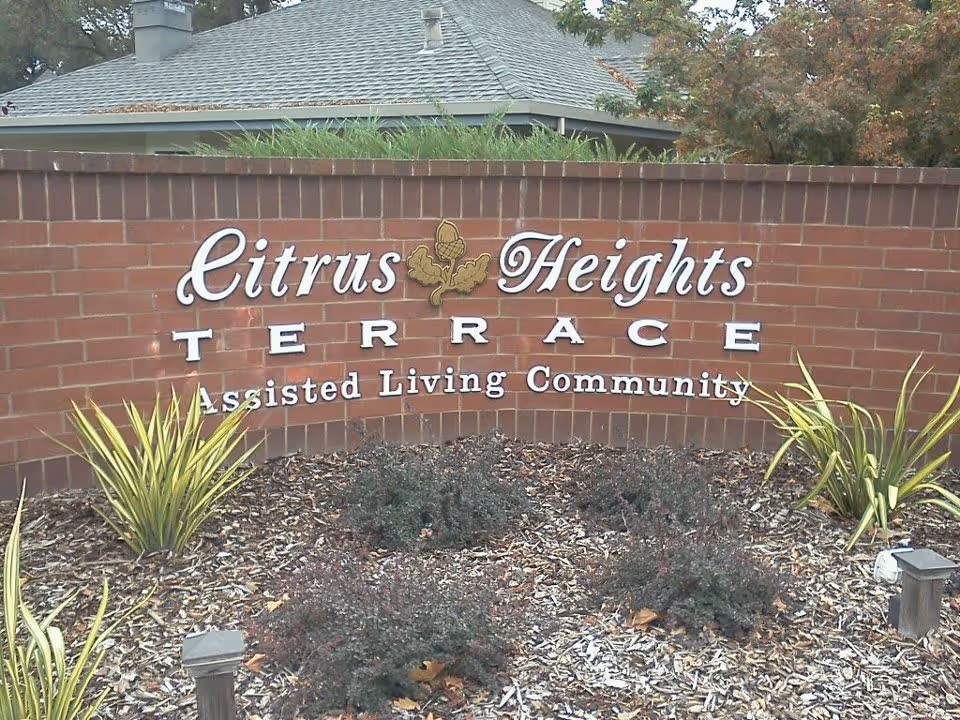 Brick wall sign for Citrus Heights Terrace Assisted Living Community with plants and mulch in front and a building roof visible in the background.