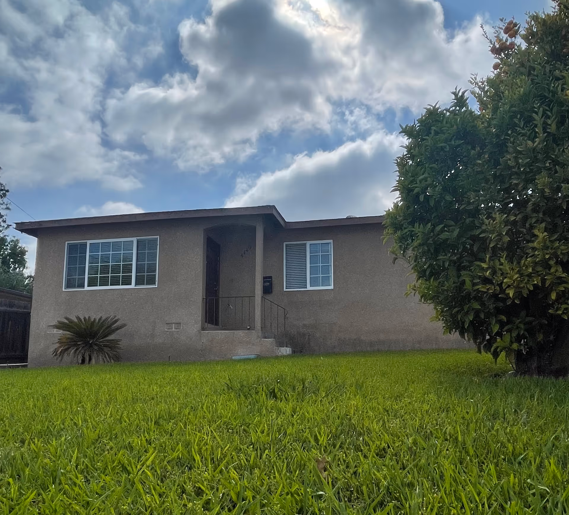 Single story beige building with front door, windows, green lawn, and a cloudy sky.