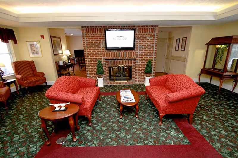 A cozy living room area with two red patterned sofas facing each other across a small wooden coffee table with magazines. The room features a green and red patterned carpet, a brick fireplace with a TV mounted above it, two small decorative topiary plants on either side of the fireplace, and additional seating including a striped armchair and a wooden side table with a lamp. There is also a wooden console table with a mirror on the right side of the room.