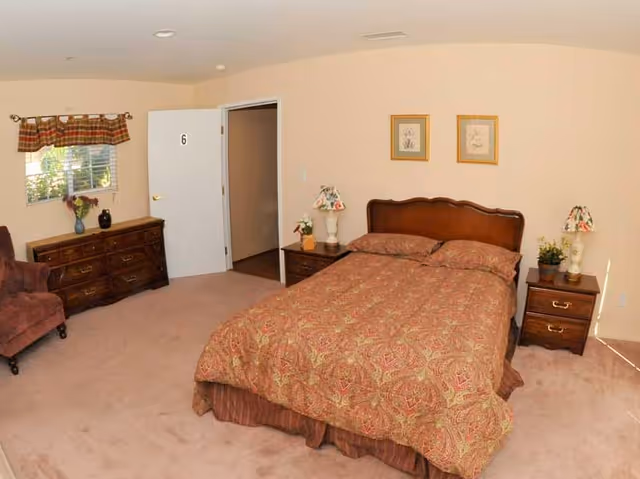 A furnished bedroom with bed, dressers, lamps, and a window in Magnolia Gardens senior facility.