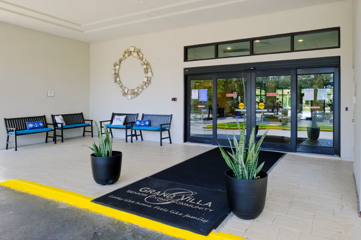 Entrance area of Grand Villa of Boynton Beach senior living community featuring automatic glass doors, a black mat with the facility's name and slogan, two large potted plants, and several black benches with blue cushions along the wall.