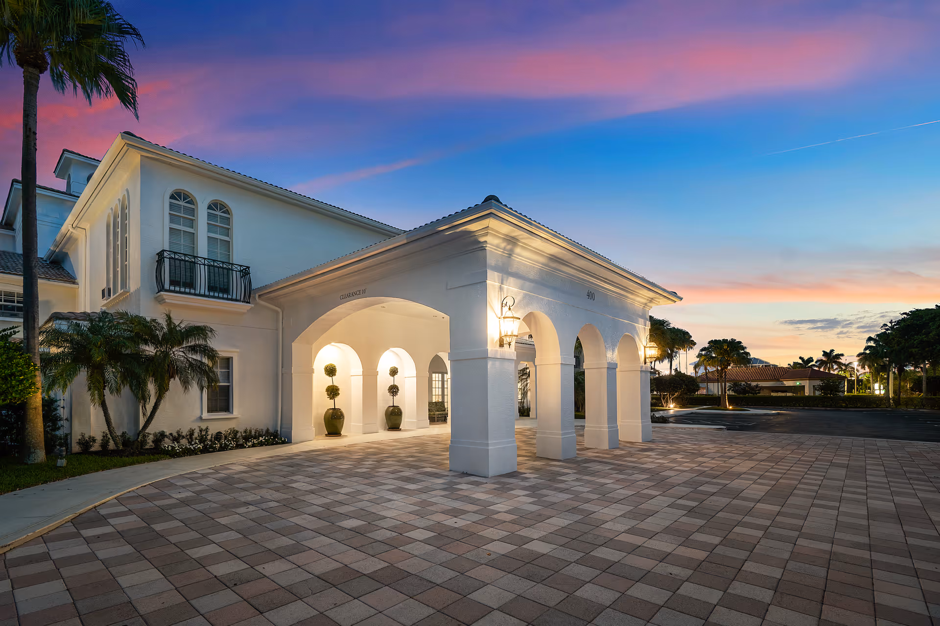White Mediterranean-style senior living facility entrance with arches and palm trees at sunset.