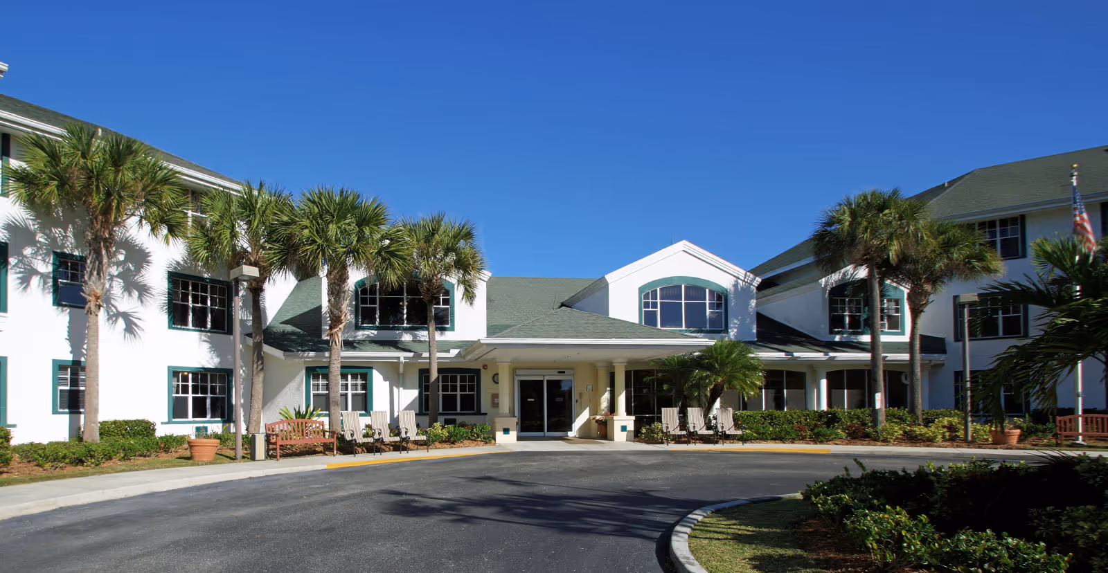 Front entrance of a white multi-story senior living building with palm trees, benches, and a circular driveway under a clear blue sky.