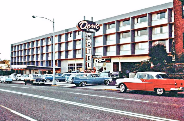 Exterior view of the Doric Tacoma Motel, a mid-century modern style building with multiple floors and rows of windows. Several vintage cars are parked along the street and in the motel parking lot. A large vertical sign reads 'Doric Tacoma Motel' with an additional smaller sign for a restaurant.