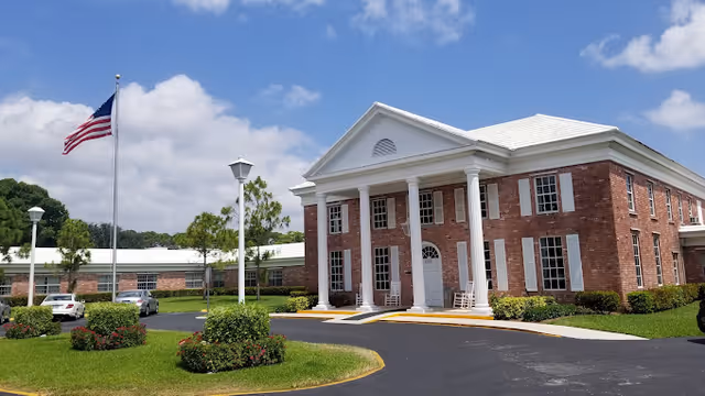 Brick senior living facility entrance with white columns, a circular driveway, landscaped grounds, and an American flag flying nearby.