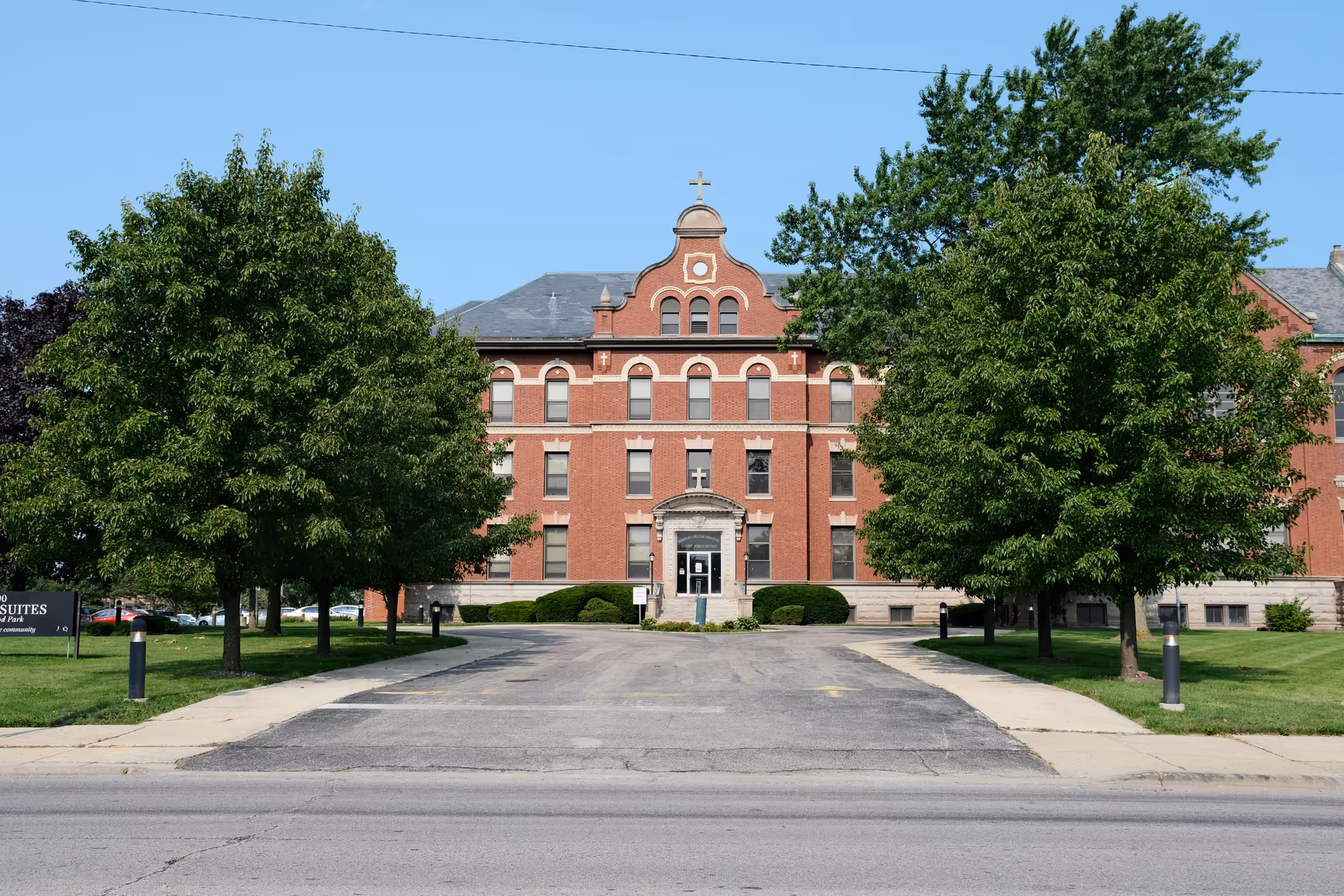Front exterior view of a large, historic red brick building with a decorative gable and a cross on top. The building is surrounded by green trees and a driveway leading to the main entrance. A sign on the left side reads 'Senior Suites'.
