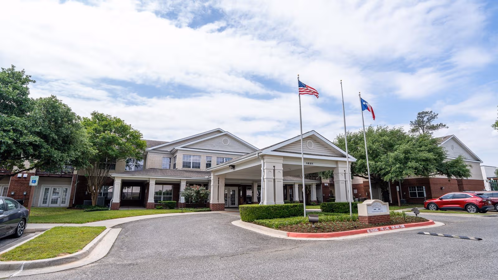 A senior living facility exterior with a covered entrance, flags, and manicured landscaping.