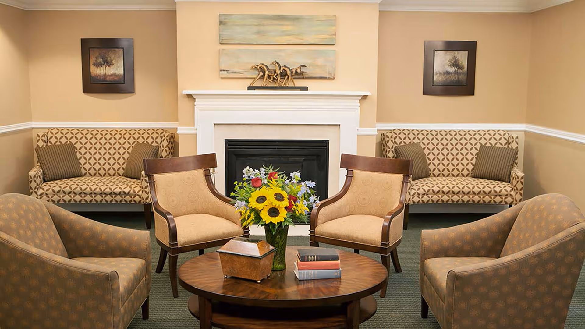 A cozy living room area with a white fireplace in the center, flanked by two patterned sofas with striped pillows. In front of the fireplace are four upholstered armchairs arranged around a round wooden coffee table that holds a vase of colorful flowers, a decorative box, and a stack of books. Two framed paintings hang on the beige walls on either side of the fireplace.