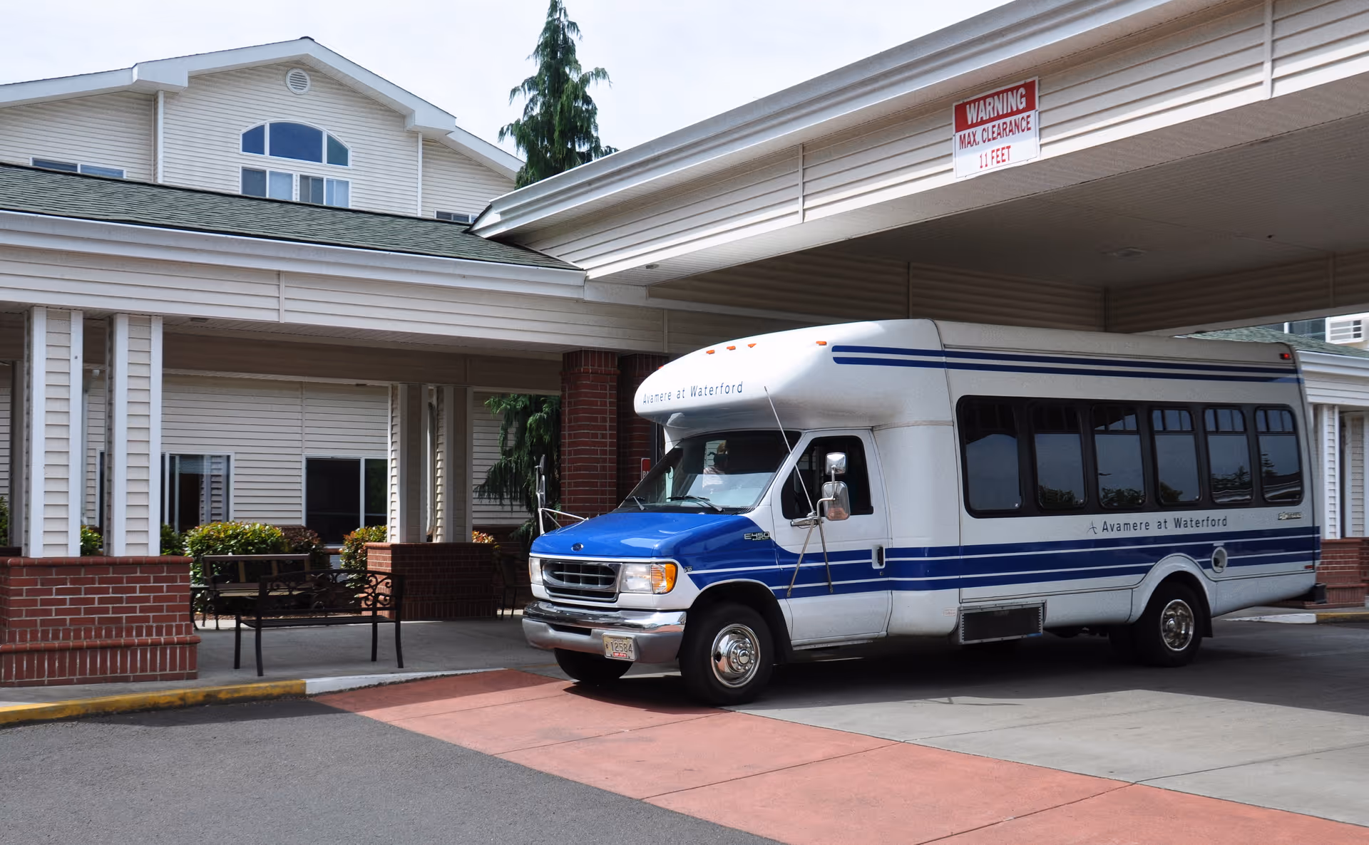 A white and blue shuttle bus parked under a covered driveway at a senior living facility. The building has beige siding with white trim and red brick accents. There is a bench and some greenery near the entrance. A sign above the driveway indicates a maximum clearance of 11 feet.