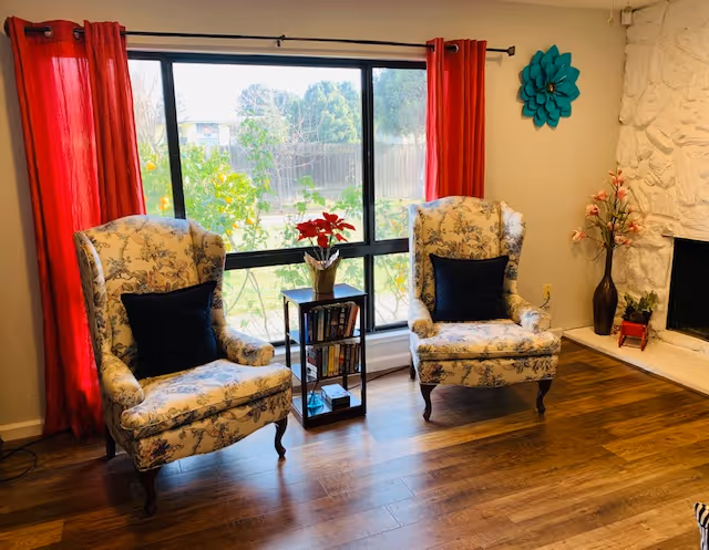 A cozy living room area with two floral upholstered armchairs each with a dark cushion, positioned on a wooden floor in front of a large window with red curtains. Between the chairs is a small black bookshelf with books and a vase with red flowers on top. To the right is a white stone fireplace with decorative vases and flowers.