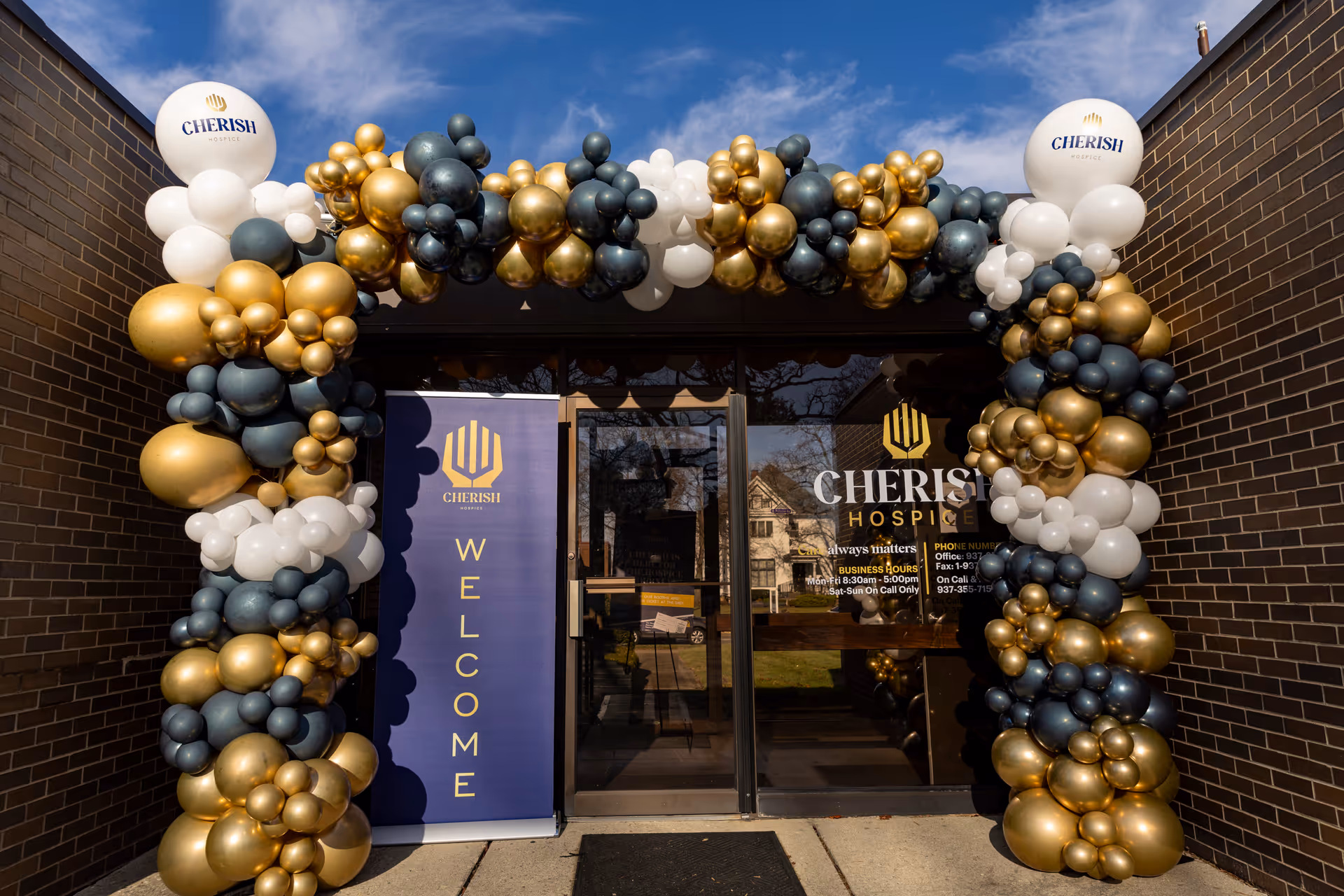 Entrance to Cherish Hospice building adorned with gold, navy, and white balloon arch.