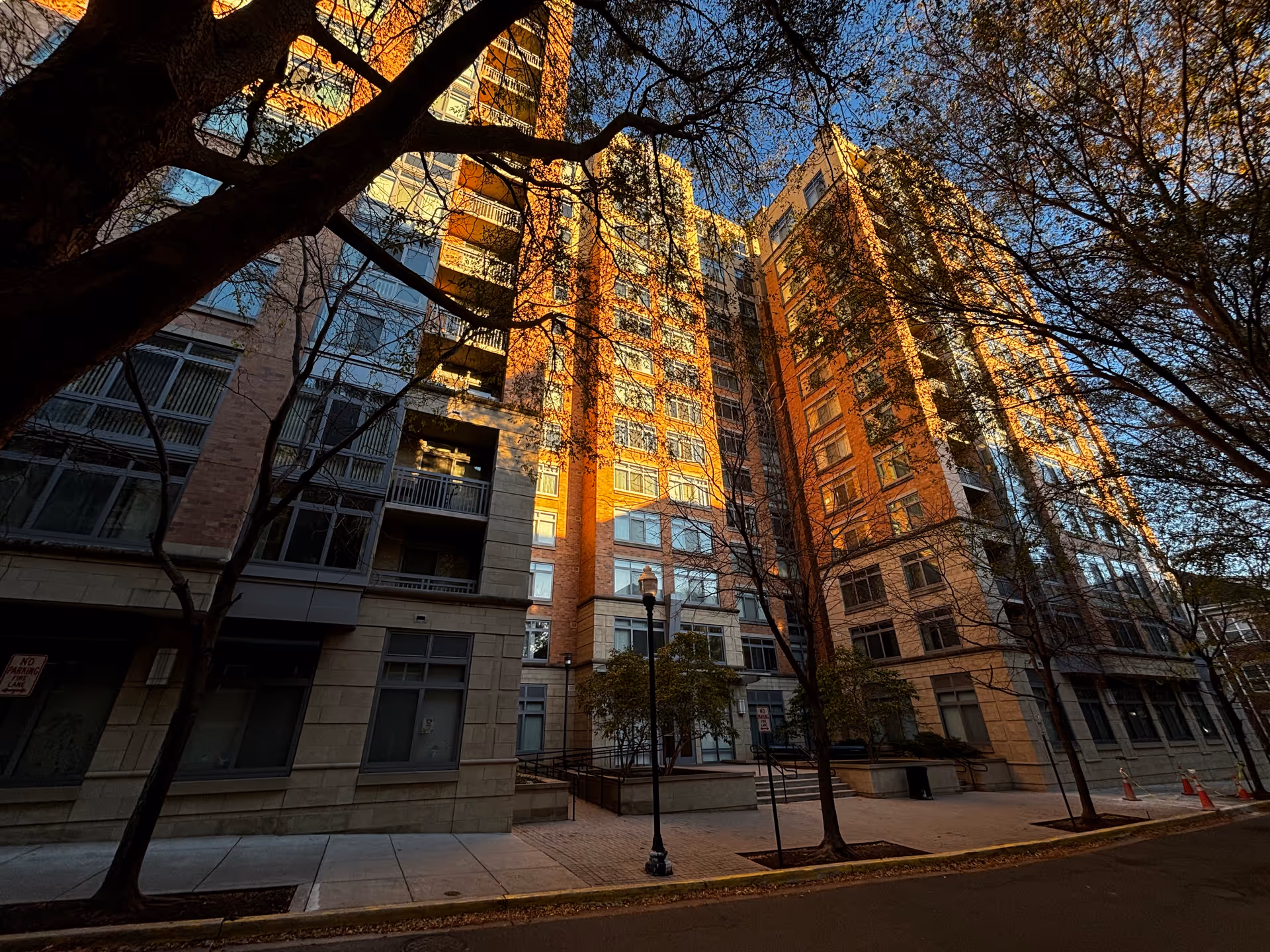 Low-angle view of a multi-story brick-and-glass residential building lit by warm sunset light, with trees and a sidewalk in front.