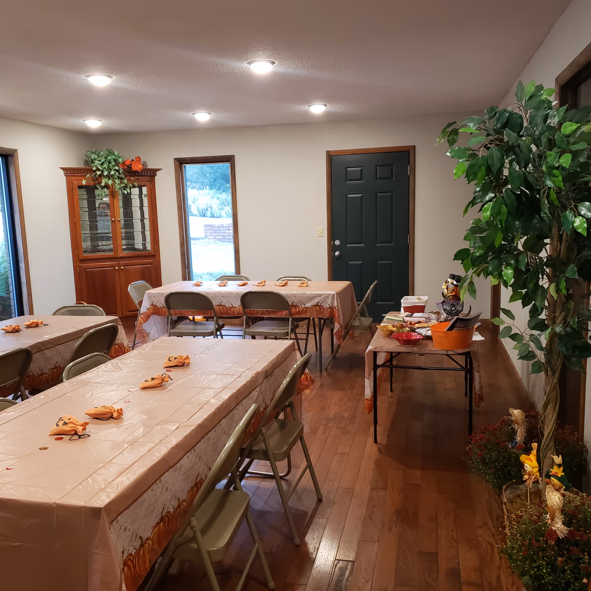 Indoor senior living dining room with long tables, folded napkins, and potted plants.