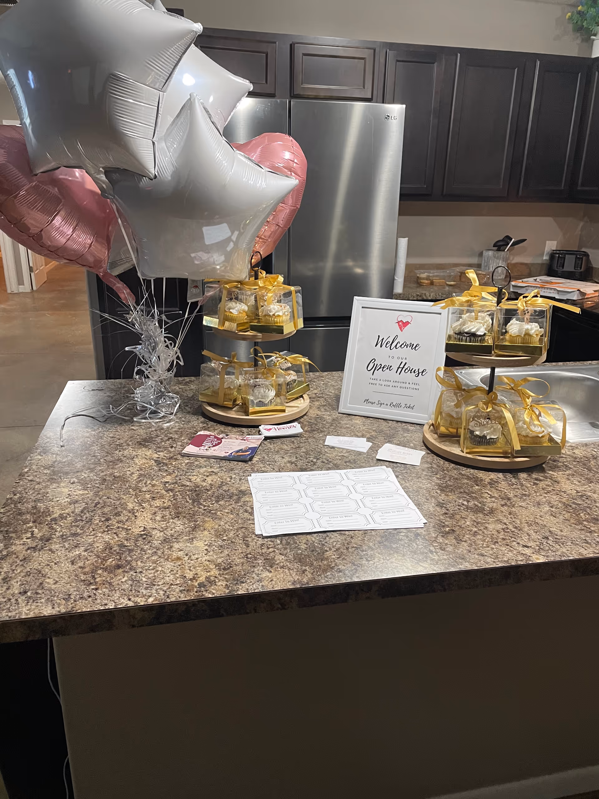Kitchen island decorated for an open house with tiered trays of cupcakes, balloons, and a welcome sign in front of dark cabinets and a stainless refrigerator.