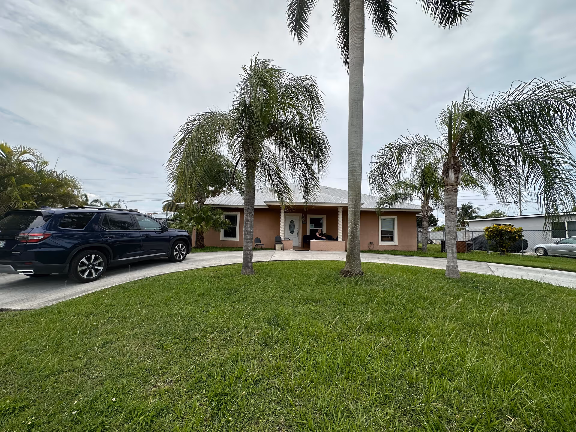 Single-story brick senior living facility with driveway, palm trees, and a parked SUV at front.