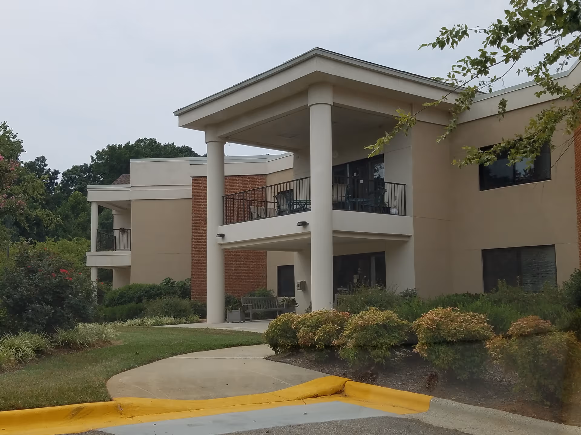 Two-story beige senior living building with balconies, landscaping, and a curved walkway at the entrance.