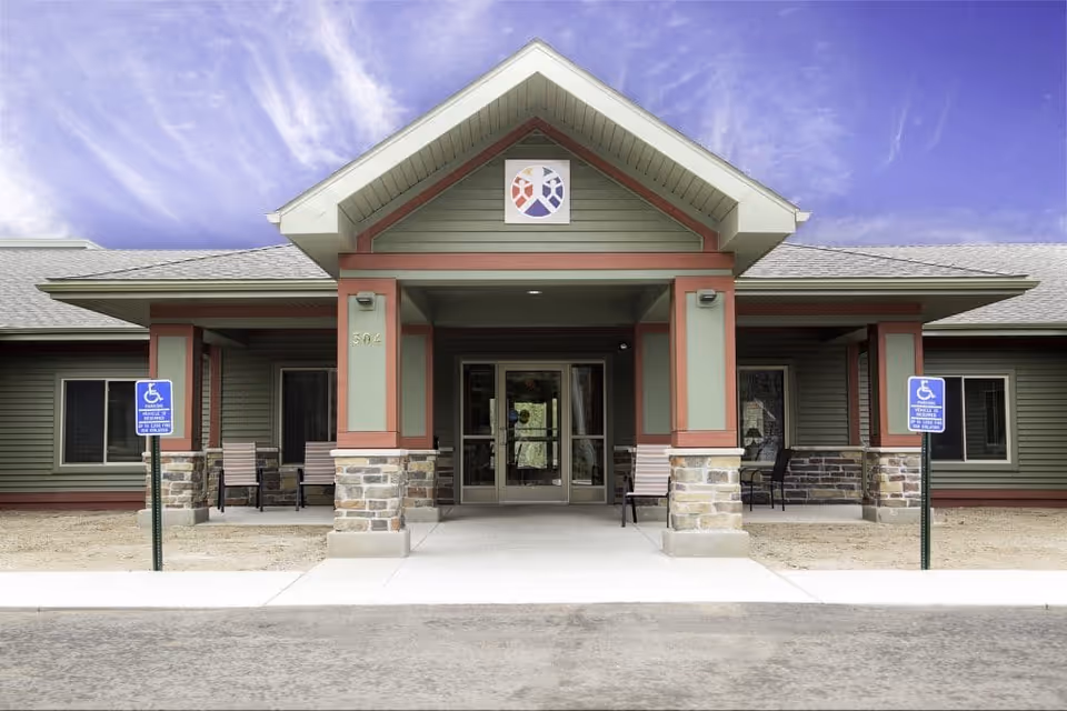Front exterior view of a single-story building with a covered entrance supported by stone and wood pillars. The building has green siding with red trim and a gray shingled roof. There are two handicap parking signs in front of the entrance and several chairs placed on the porch area. The sky is partly cloudy with a blue hue.