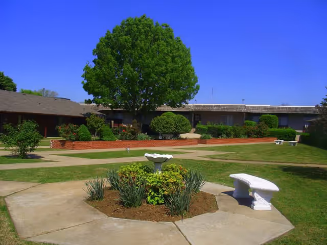 Sunny outdoor courtyard of Southern Hills Rehabilitation Center with trees, benches, and flower beds.