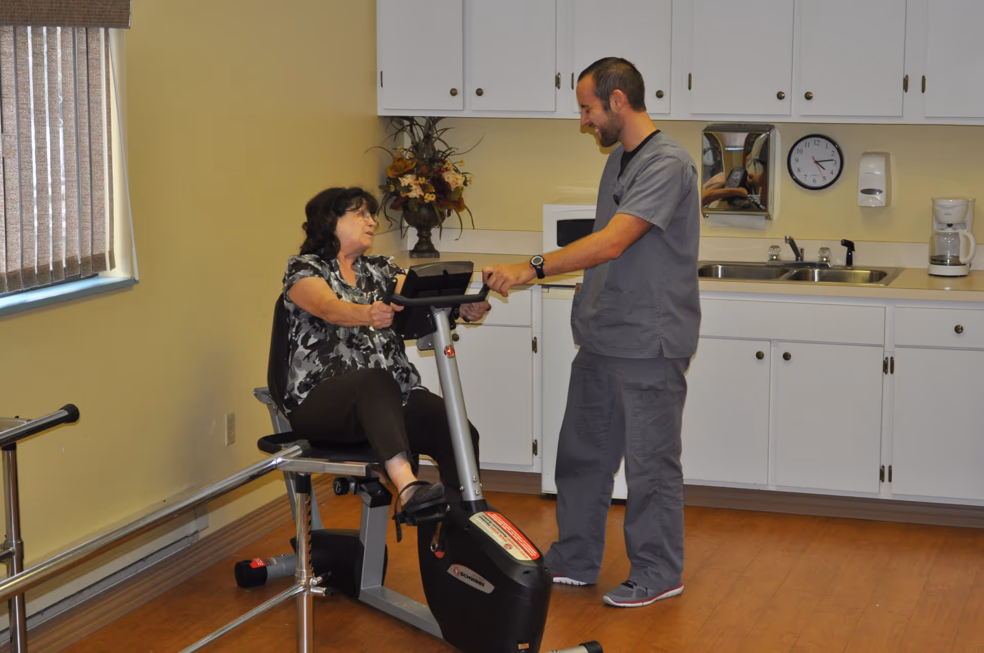 A woman is seated on a stationary exercise bike in a room with wooden flooring and white cabinets. A man in gray scrubs is standing next to her, smiling and holding the bike's handlebars, assisting or encouraging her. The room has a window with vertical blinds, a clock on the wall, a coffee maker, and a decorative flower arrangement on the counter.
