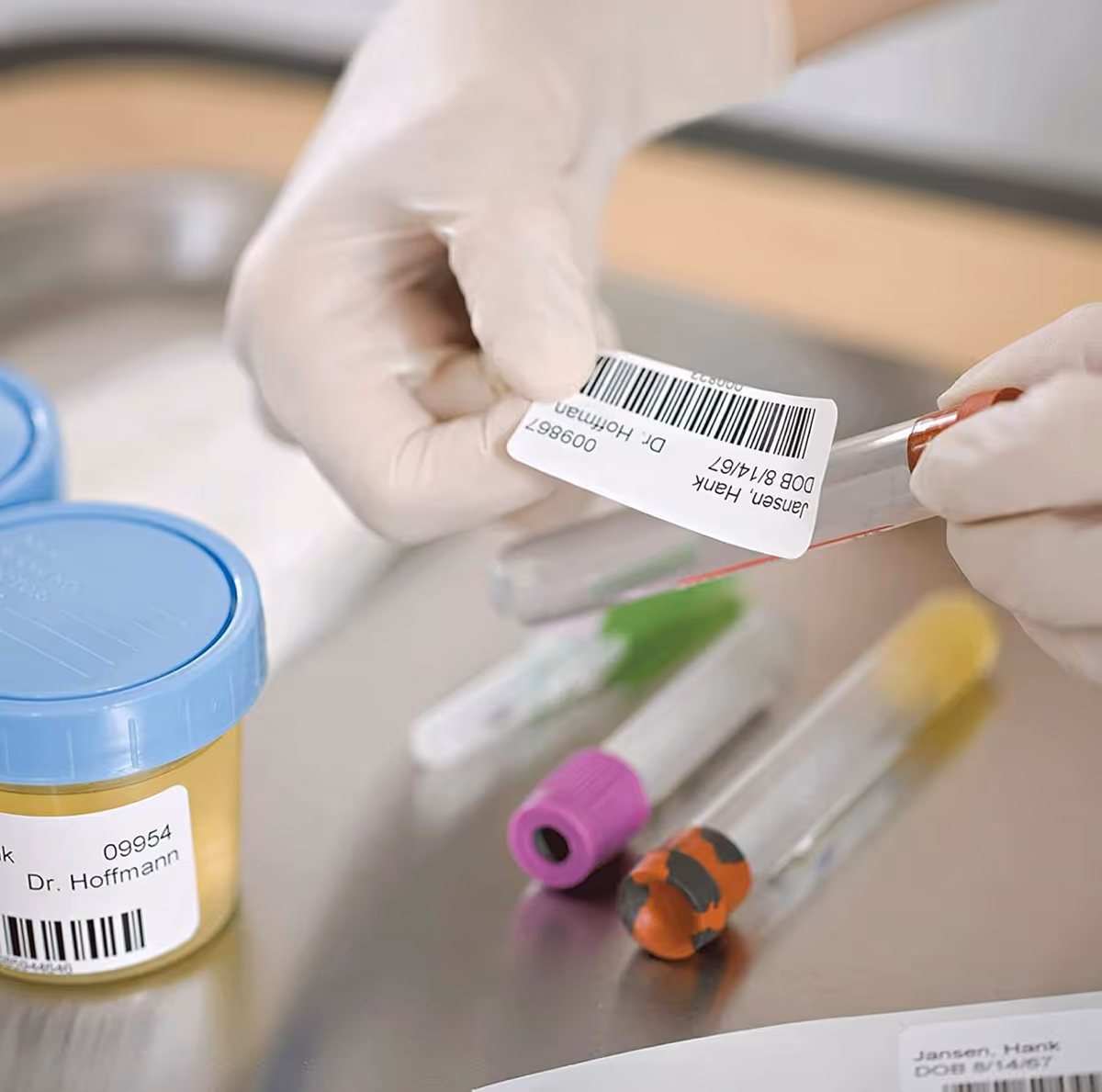 Gloved hands attach a barcode tag to test tubes on a stainless lab bench.