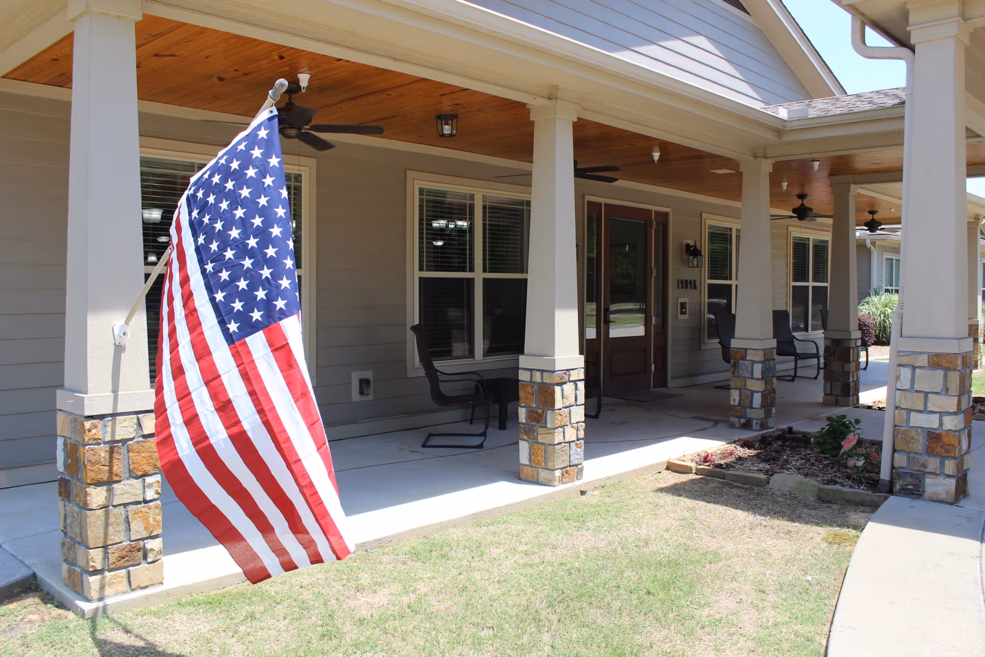 Front porch of a senior living facility with stone pillars, a wooden ceiling with ceiling fans, black chairs, and an American flag mounted on one of the pillars. The entrance door has a glass panel with the words 'Home Sweet Home' visible.