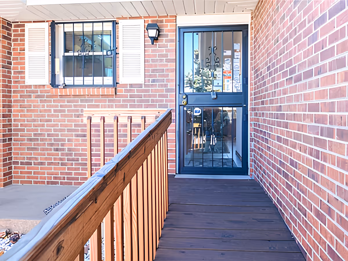 A brick exterior hallway leading to a black metal security door with glass panels. To the left of the door is a window with white shutters and black security bars. A wooden railing runs along the left side of the hallway.