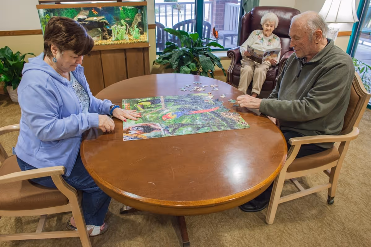 Two elderly individuals sitting at a round wooden table working on a colorful jigsaw puzzle, while another elderly person sits in a leather armchair in the background reading a magazine. The room has a large fish tank, green plants, and windows showing an outdoor patio area.