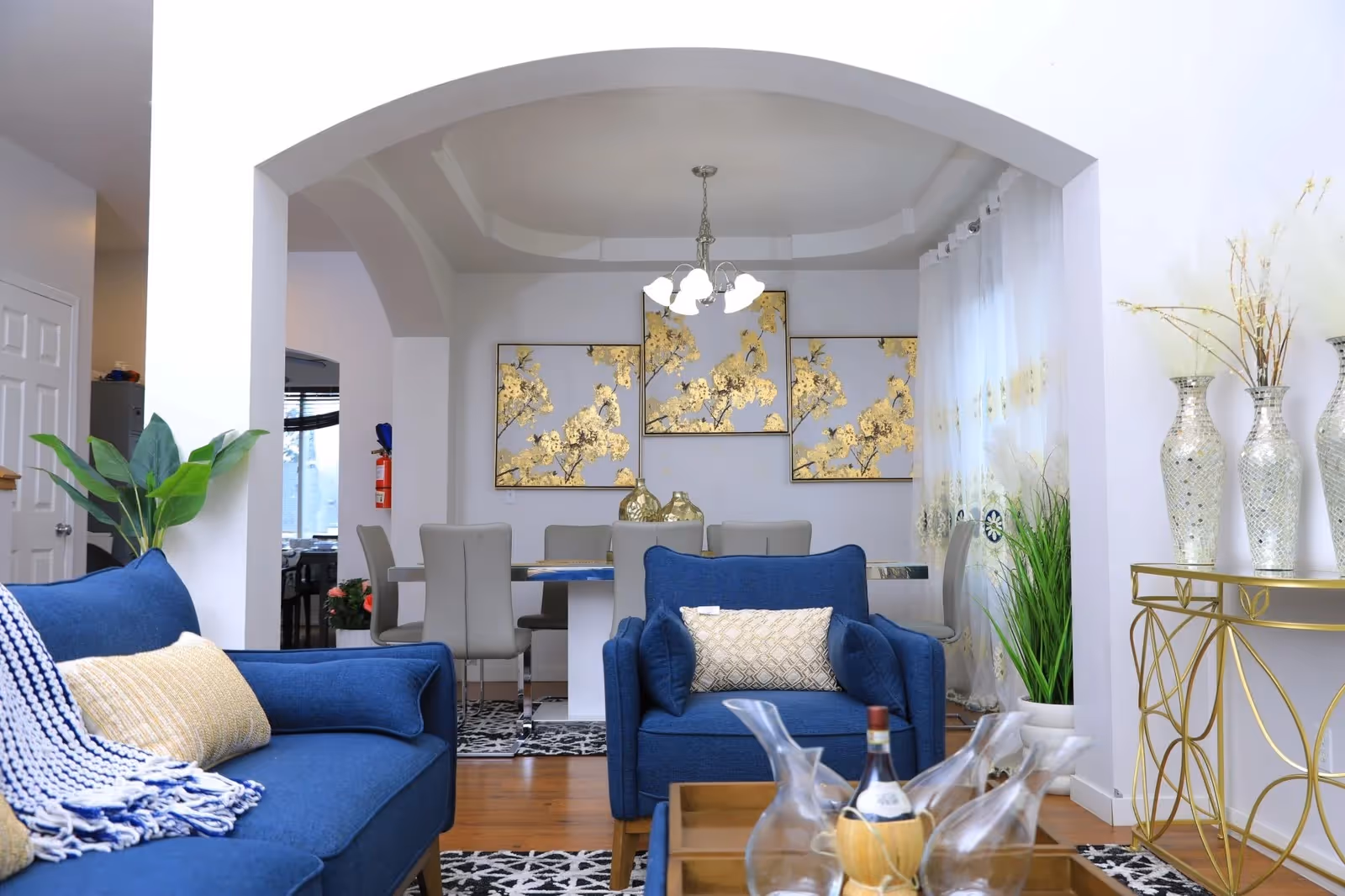 Bright living room in a senior facility with blue sofas, gold decor, and dining area beyond.
