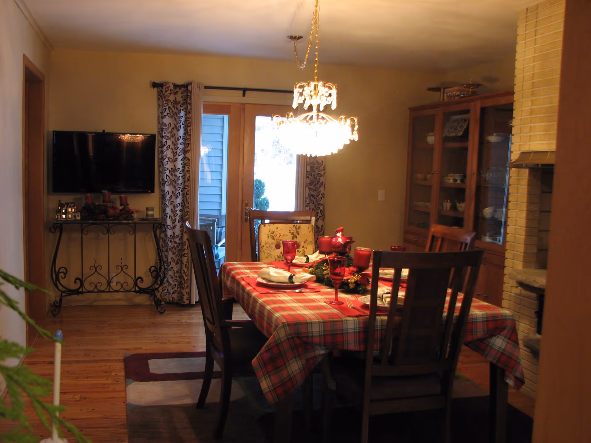 Dining room with a table set using a red plaid tablecloth, wooden chairs, a chandelier overhead and a china cabinet.