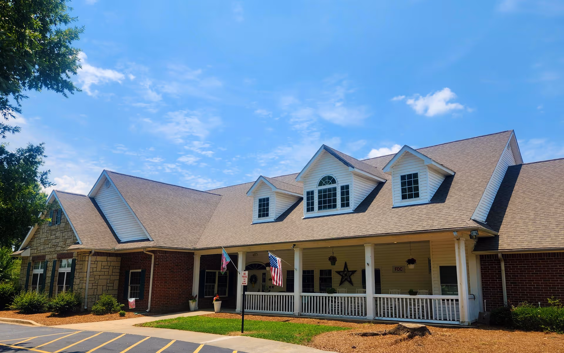 Exterior view of a single-story senior living facility building with a combination of brick and stone walls, a beige shingled roof with dormer windows, a covered porch with white railings, and two flags displayed near the entrance under a partly cloudy blue sky.